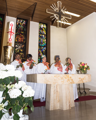 Die fünf Frauen des Gospelchors in weißen Gewändern und orangefarbenen Halstüchern, stehend hinter dem Marmoraltar und dem von der Decke herabhängenden Kreuz. Seitlich befindet sich die Osterkerze und Blumenschmuck.