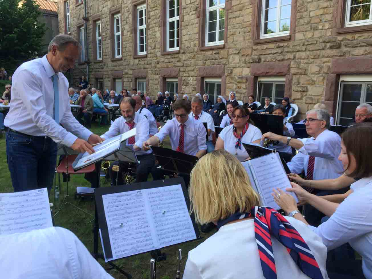 Im Vordergrund die Musiker der Stadtkapelle Heppenheim in weißen Hemden und Blusen, die Männer mit rotblauen Krawatten, die Frauen mit rotblauen Halstüchern. Im Hintergrund sitzen die Zuhörer auf Bänken und Stühlen vor dem Westflügel des Klostergebäudes.