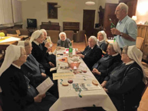 Die Schwestern des Sankt Vinzenklosters in fröhlicher Runde während der Weinprobe im Refektorium des Mutterhauses. Zum Wein werden Wasser und Brot gereicht, das sich auf dem Tisch befindet, andem die Schwestern sitzen. Rechts im Bild steht der Winzer Hans Engelhard mit einer Flasche Wein.