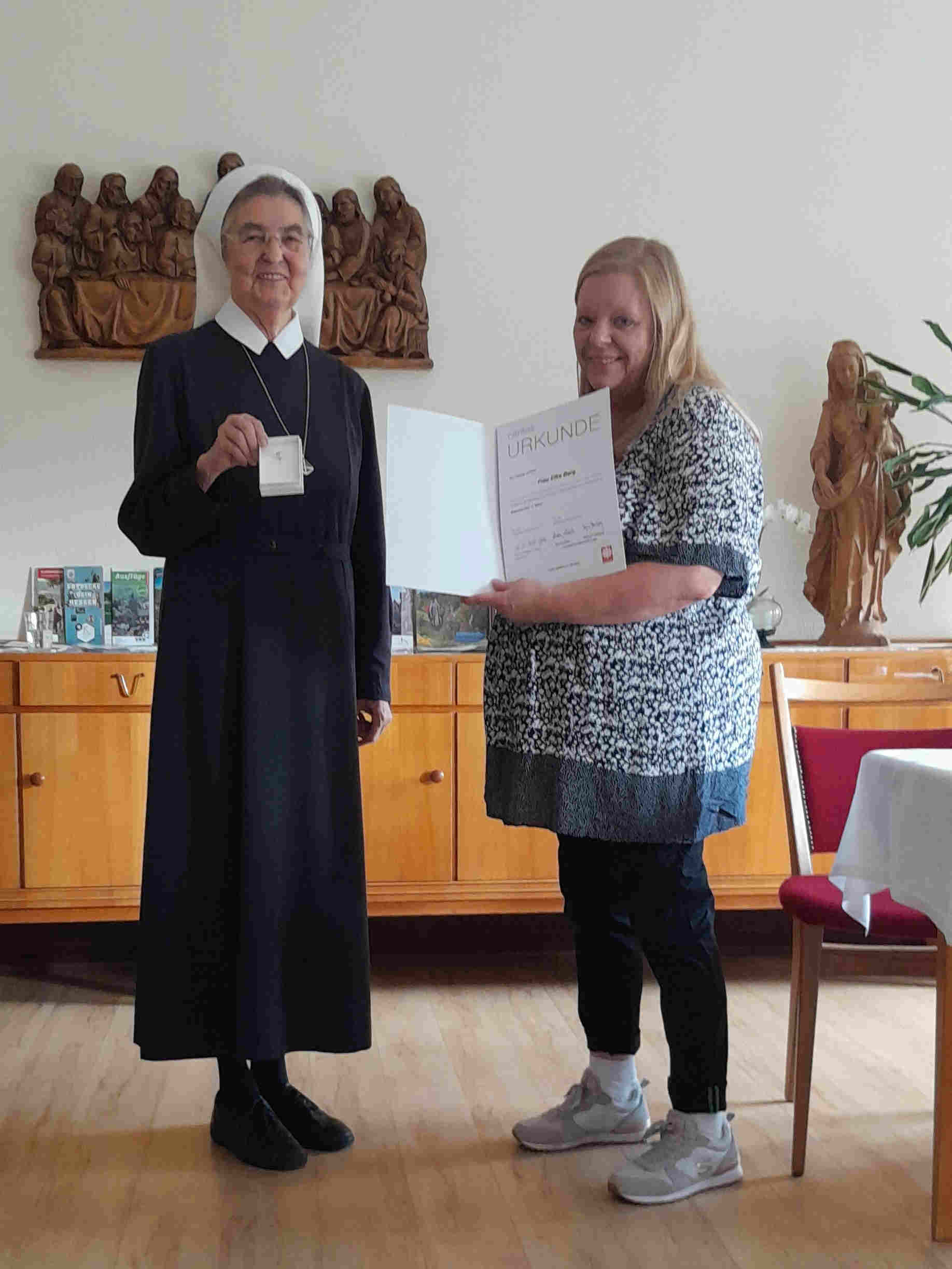 Elke Berg zeigt die Urkunde, die sie von Schwester Brigitta zum 15. Dienstjubiläum erhalten hat. Schwester Brigitta zeigt die silberne Ehrennadel, die vom Caritasverband vergeben wird. Im Hintergrund steht ein buchefarbenes Sideboard mit Prospekten darauf, sowie eine Holzstatue der Mutter Gottes mit dem Jesuskind. An der Wand darüber hängt ein Holzschnitt der das letzte Abendmahl zeigt.